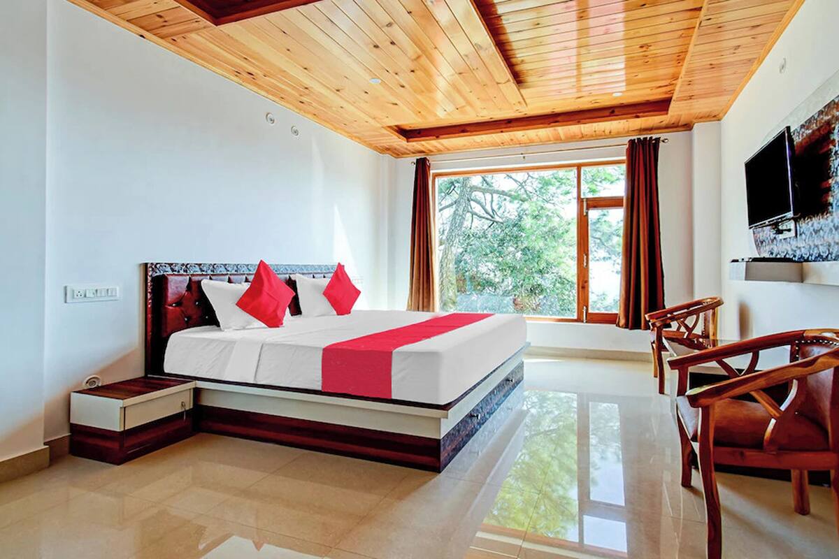 A bedroom with a wooden ceiling, a large window with curtains, a bed with red pillows, a bedside table, and a TV mounted on the wall.