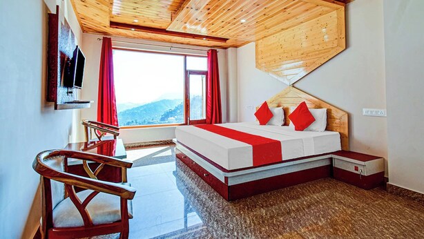 Un dormitorio con techo de madera, una cama con ropa de cama roja y blanca, un televisor montado en la pared y una ventana con vistas a las montañas.