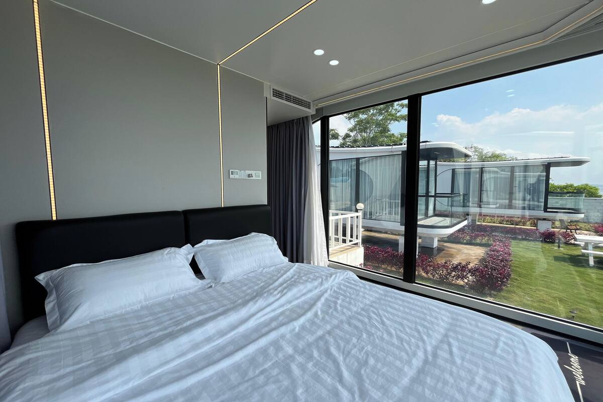 A modern hotel room with a large bed, a view of the outdoor balcony, and a glass wall.