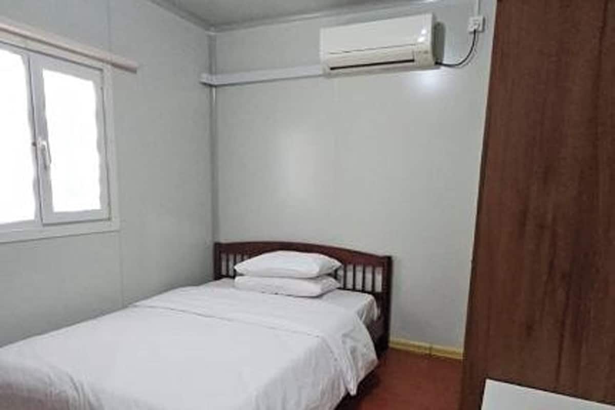A small, single-bed room with a wooden headboard, white bedding, and an air conditioning unit mounted on the wall.