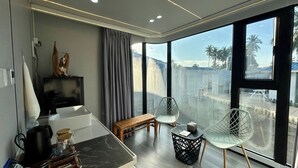 Ein modernes Zimmer mit großem Fenster, einer Holzbank, einem Metallstuhl, einem kleinen Tisch und einer Küchenzeile mit Wasserkocher und Tablett.
