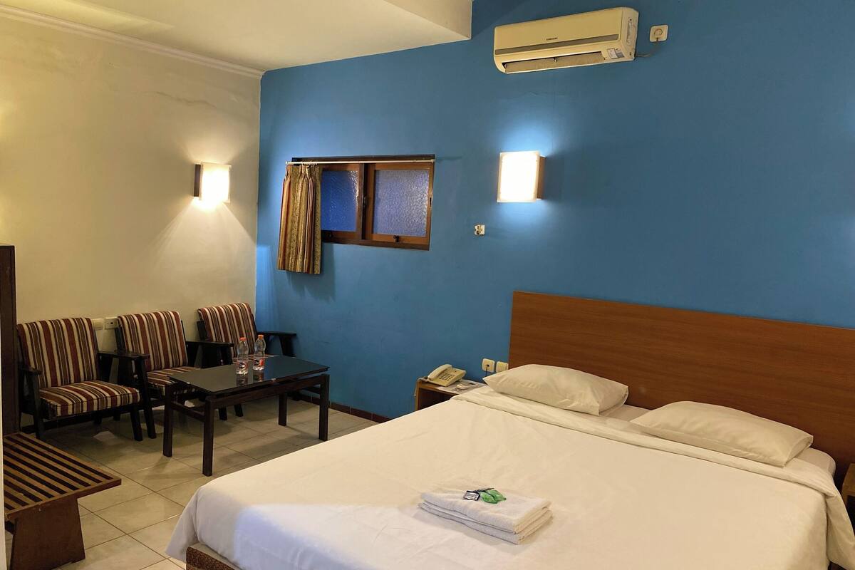 A hotel room with a large bed, a wooden headboard, a nightstand with a phone, and a window with curtains. There are two striped chairs and a small table with a bottle and glasses.