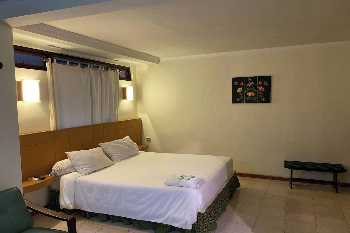 A hotel room with a bed, a bench, a wall-mounted artwork, and a window with curtains.