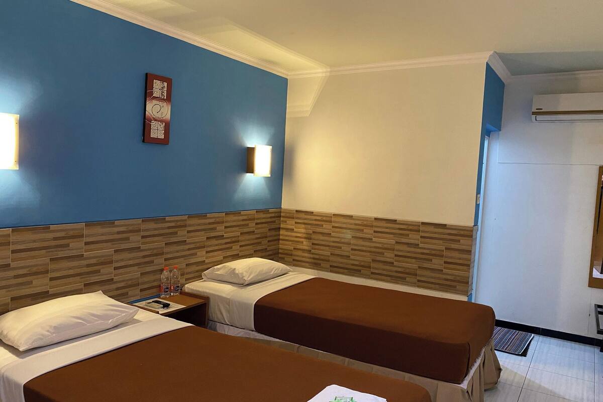 A hotel room with two beds, a wooden headboard, a wall clock, and a wall-mounted light fixture.
