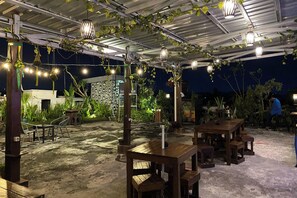 An outdoor patio area with wooden tables and chairs, string lights, and hanging plants. - Hotel Lestari (Patrang)