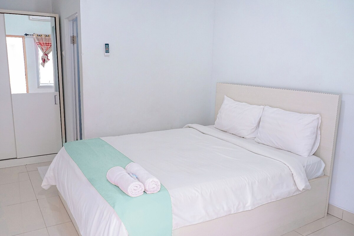 Una cama bien hecha con ropa blanca y un plaid verde claro, un armario blanco y un espejo con una toalla colgada.