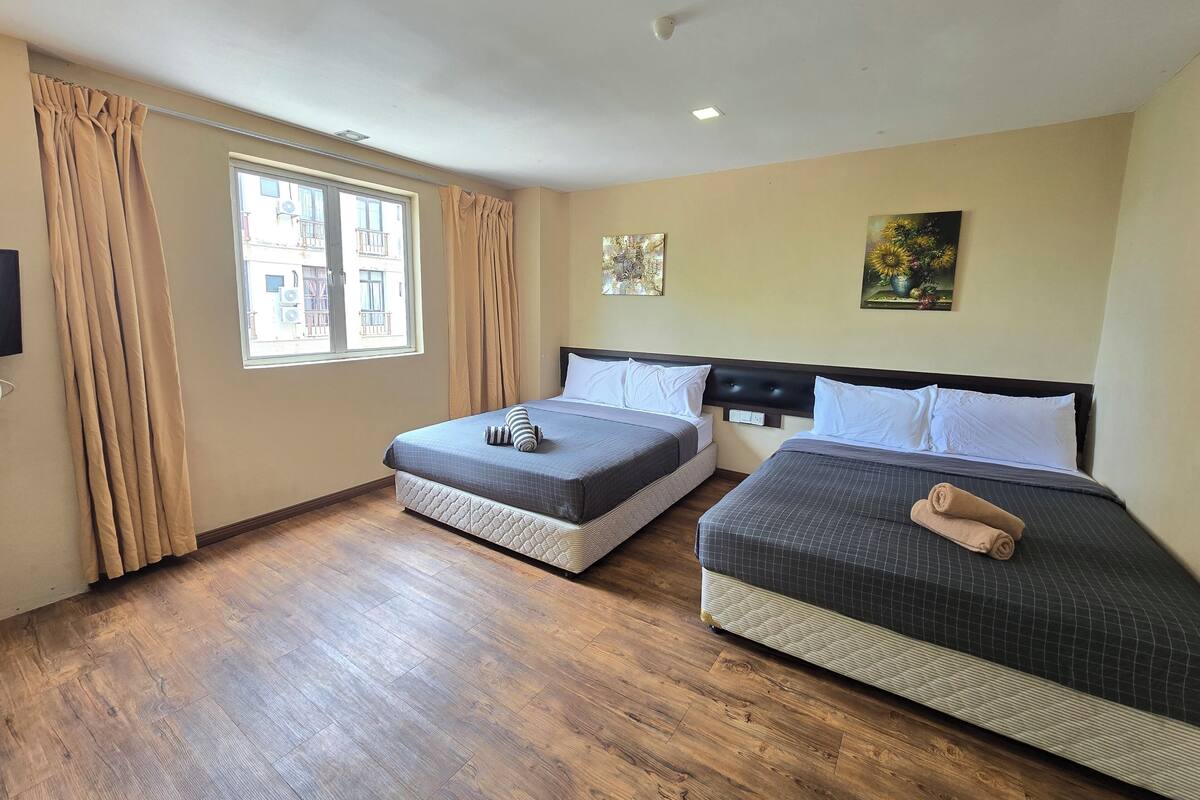 A hotel room with two beds, wooden flooring, beige walls, a window with curtains, and a ceiling light.