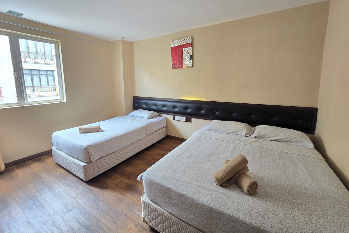 A hotel room with two single beds, a window with a view of buildings, a wall-mounted light, and a small artwork.