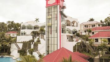 Experience the modern architecture of this resort, featuring a striking tower with a glass facade and red roof, surrounded by lush greenery and multiple buildings.