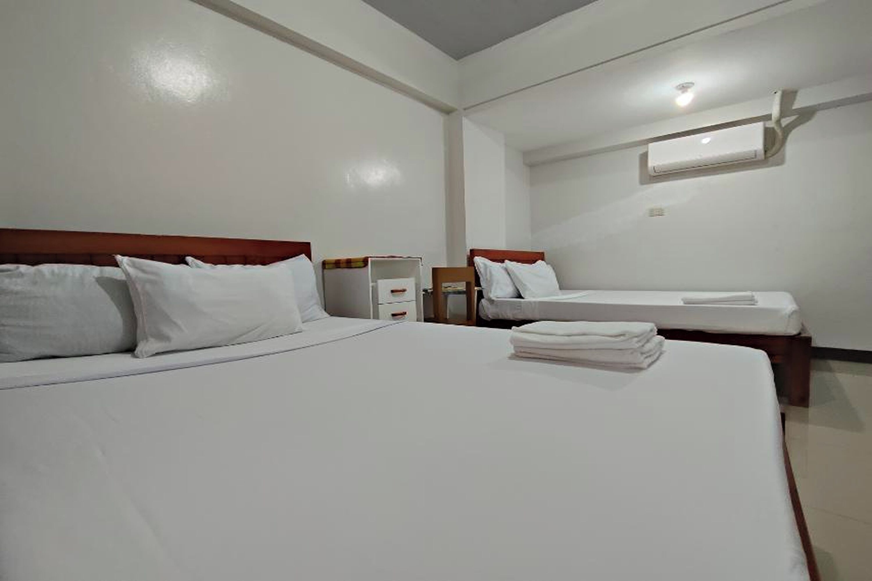 A hotel room with two beds, white bedding, a wooden headboard, a small table, and a wall-mounted air conditioning unit.