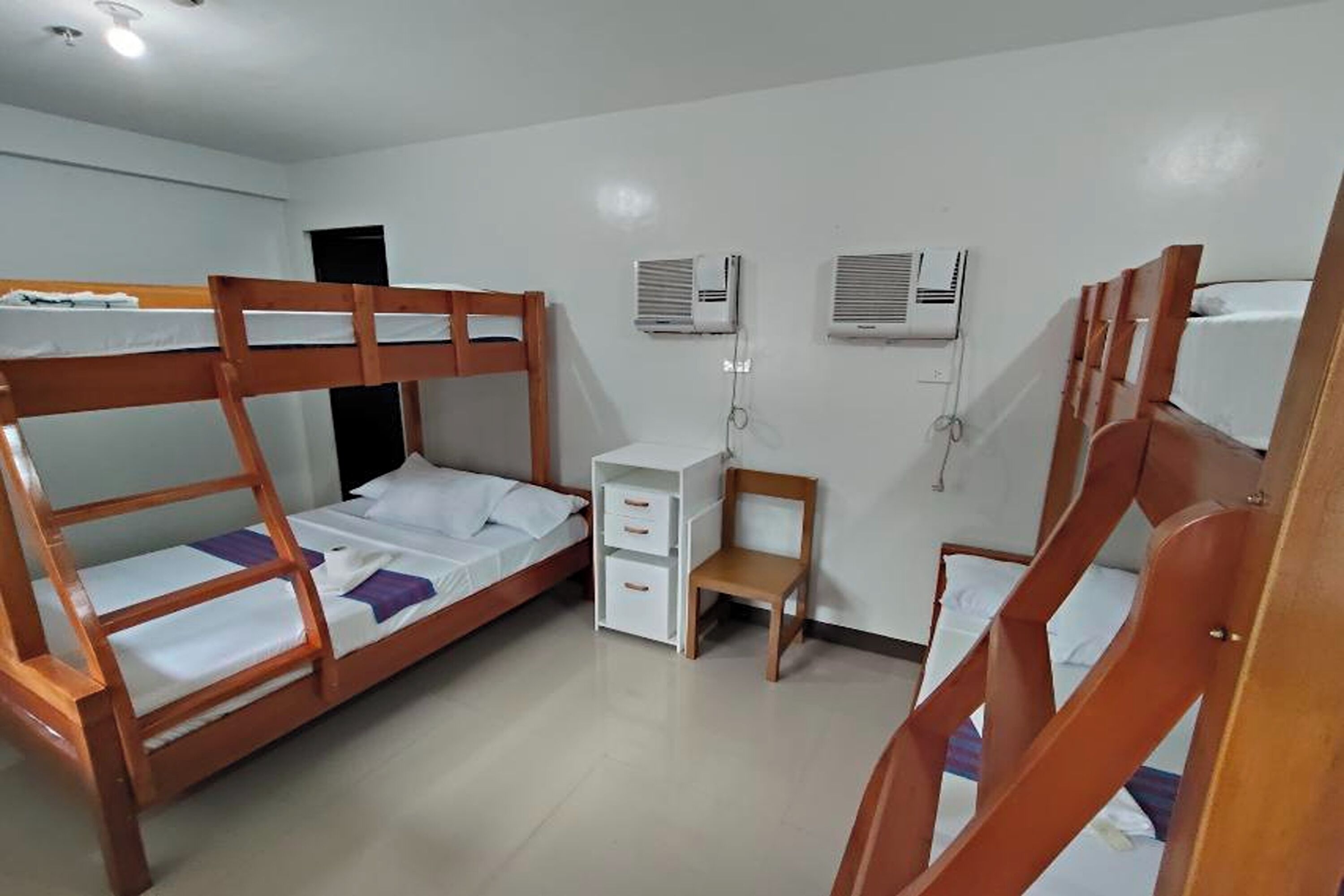 A room with two bunk beds, a small wooden chair, a white bedside table with drawers, and two wall-mounted air conditioning units.