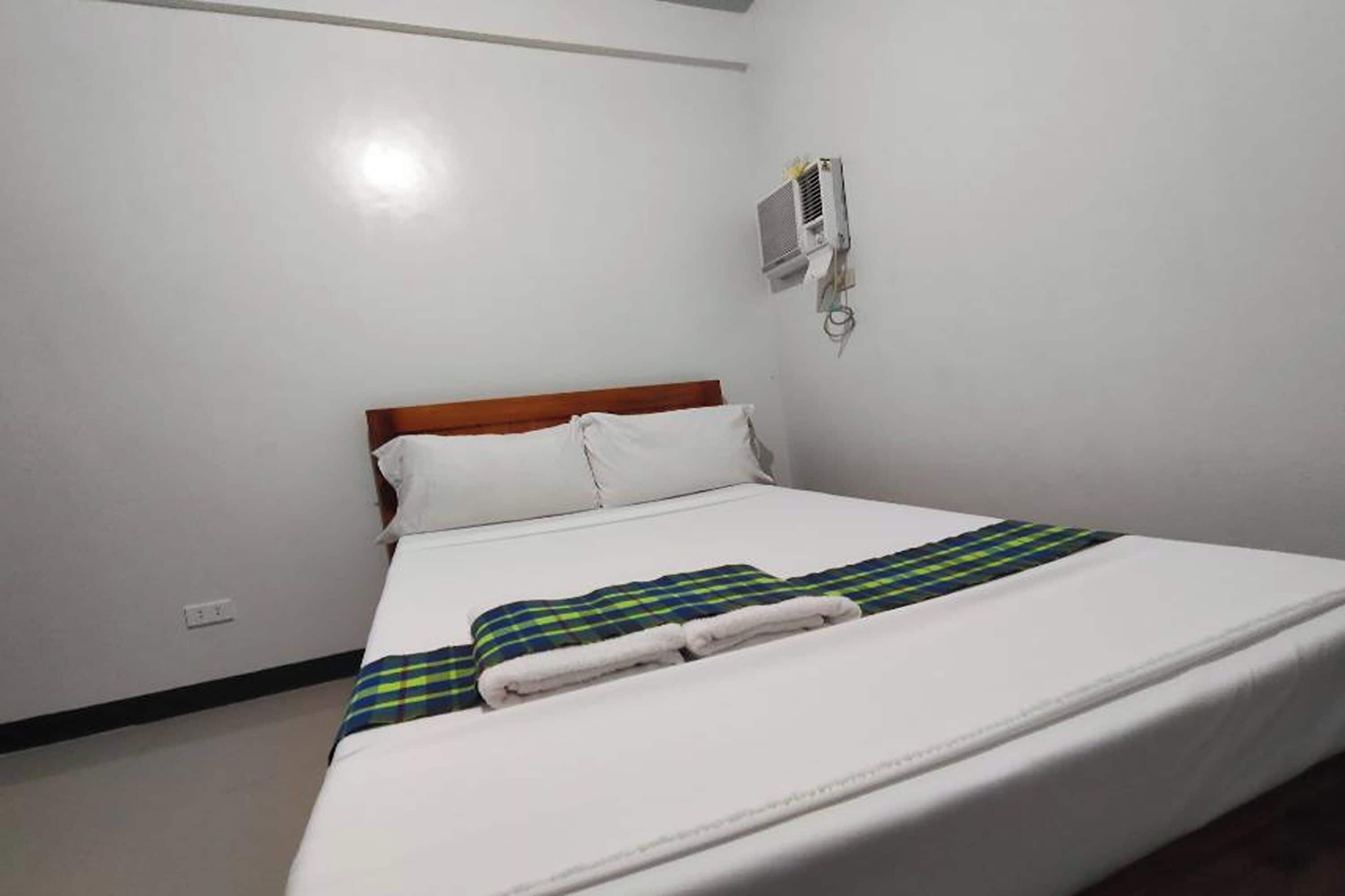 Una cama bien hecha con sábanas blancas y una colcha escocesa, un cabecero de madera y un aire acondicionado instalado en la pared.