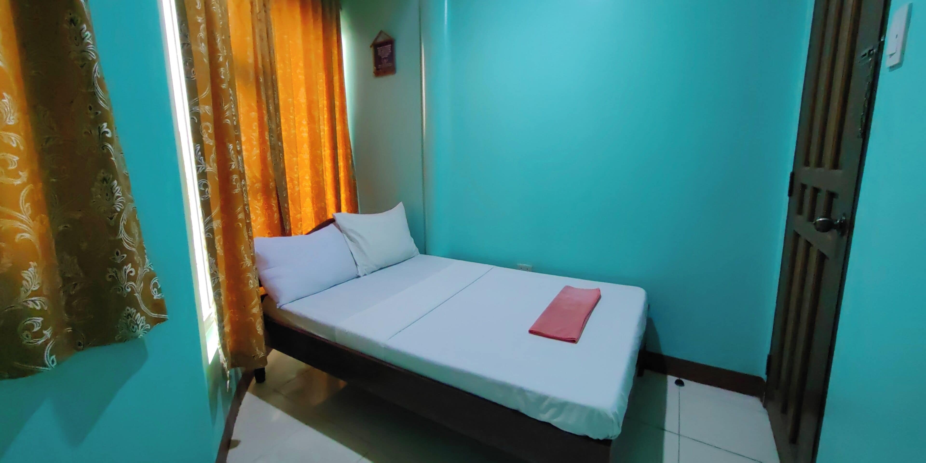 Una cama individual con colchón y almohadas blancos, una toalla roja doblada y una cortina con estampado floral.