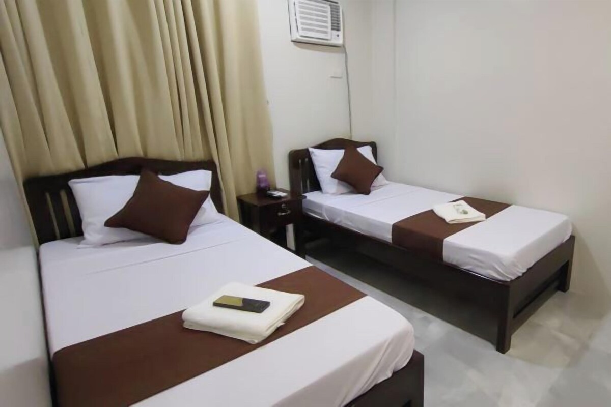 Ein Hotelzimmer mit zwei Einzelbetten, jeweils mit brauner Kopfteil und weißer Bettwäsche. Ein kleiner Nachttisch mit Lampe und Fernbedienung ist vorhanden.