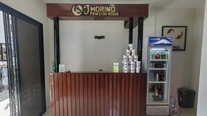 Een receptiebalie met een bordje 'JM Morino Pension House'. Er staat een koelkast vol drankjes en een prullenbak aan de rechterkant.