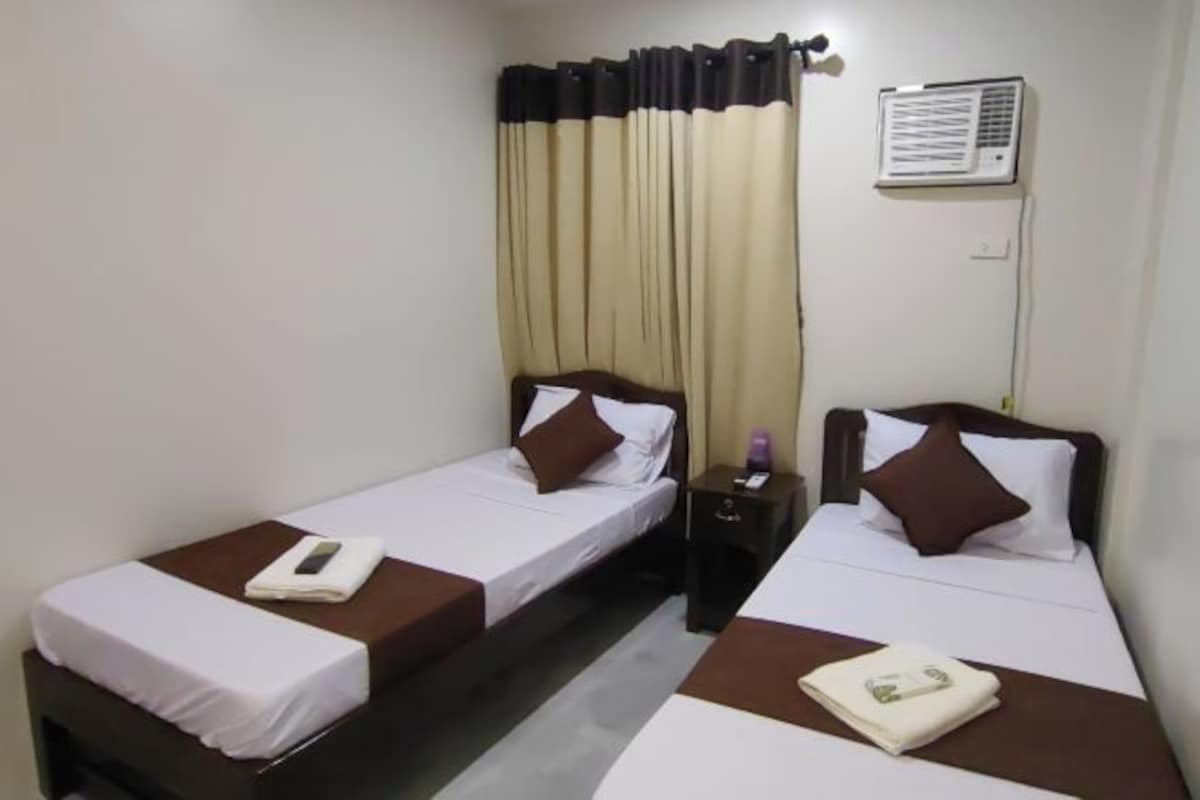 Une chambre d’hôtel avec deux lits simples, chacun équipé d’un oreiller et d’une couverture. Il y a une table de chevet avec une lampe et un téléphone sur l’un des lits, ainsi qu’un climatiseur fixé au mur.