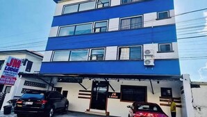 Een meerhoog gebouw met een blauw-witte gevel, een bordje met 'JM Lodge' dat een kamer te huur aangeeft, en twee auto's die ervoor geparkeerd staan.