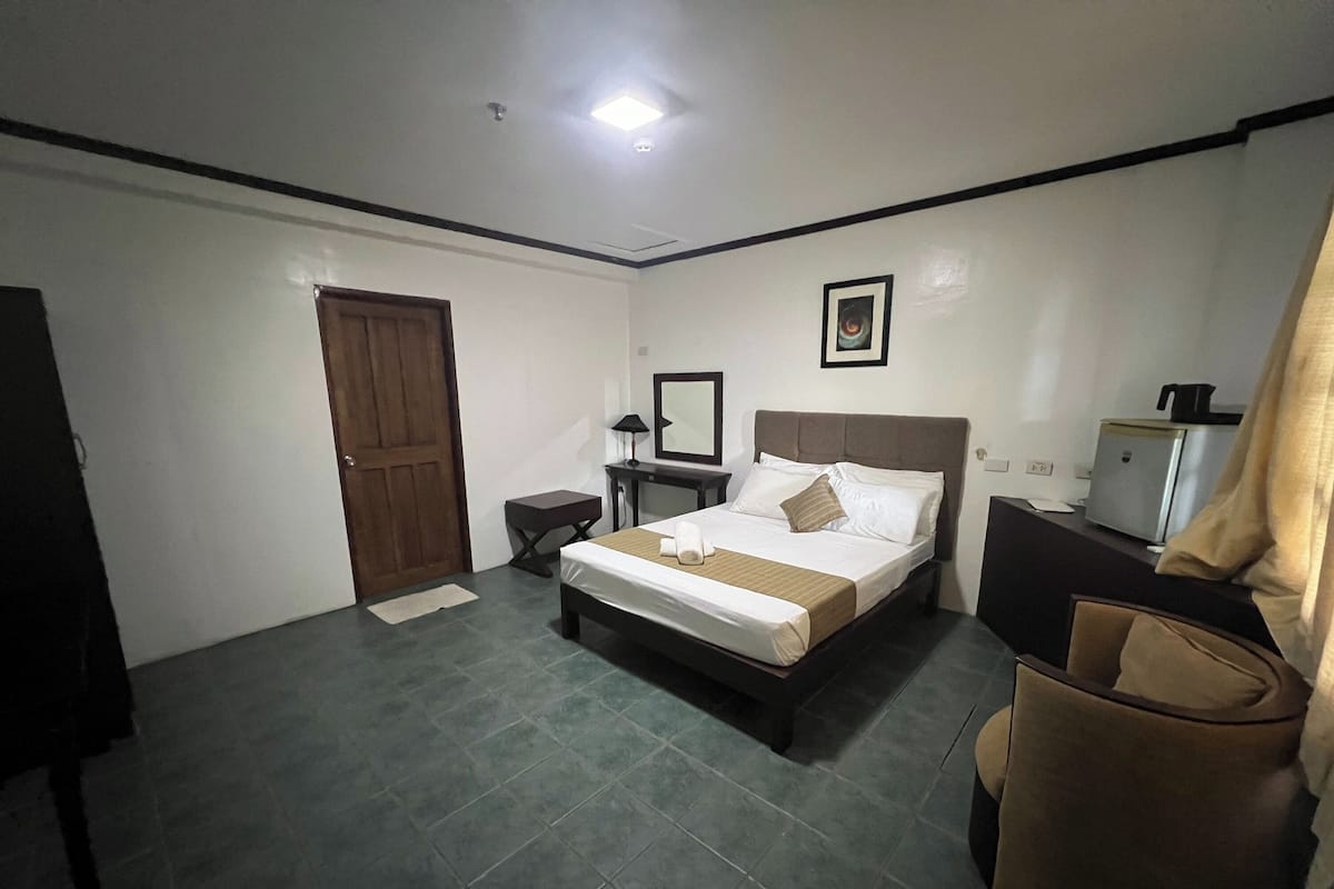 A bedroom with a bed, a nightstand, a desk, a chair, a door, and a refrigerator.