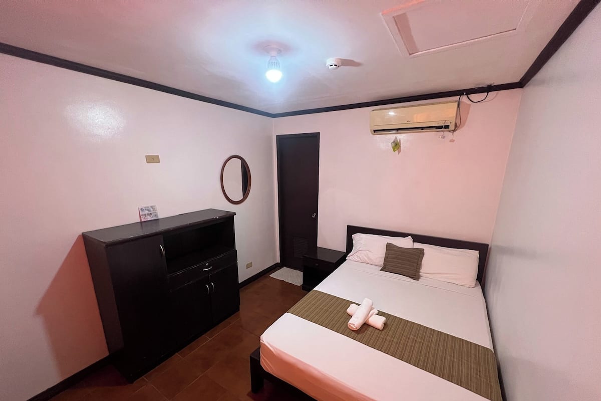 A hotel room with a bed, a desk, a mirror, an air conditioning unit, and a ceiling light.