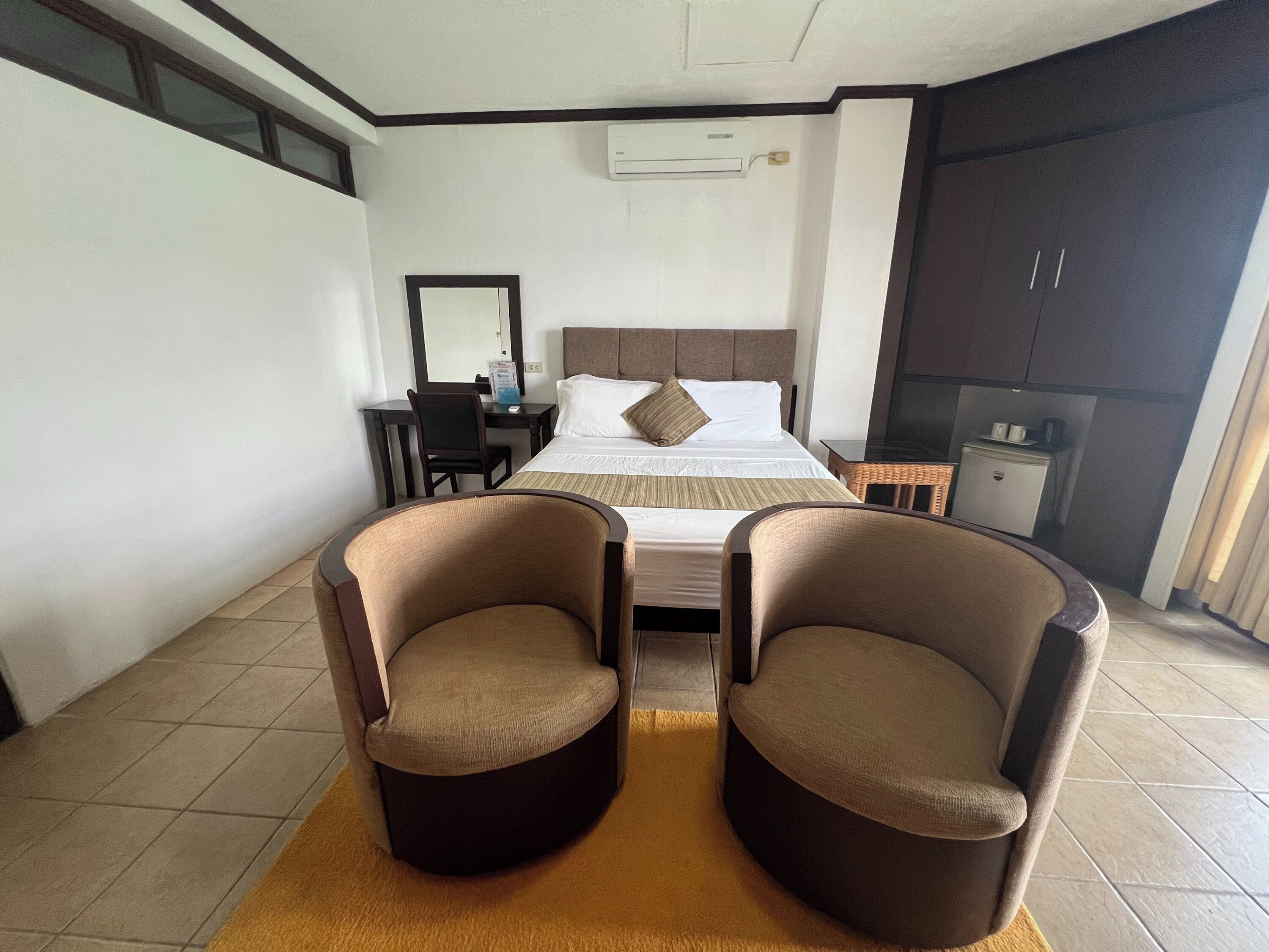 A hotel room with a bed, two armchairs, a desk, a mirror, a small kitchenette, and an air conditioning unit.
