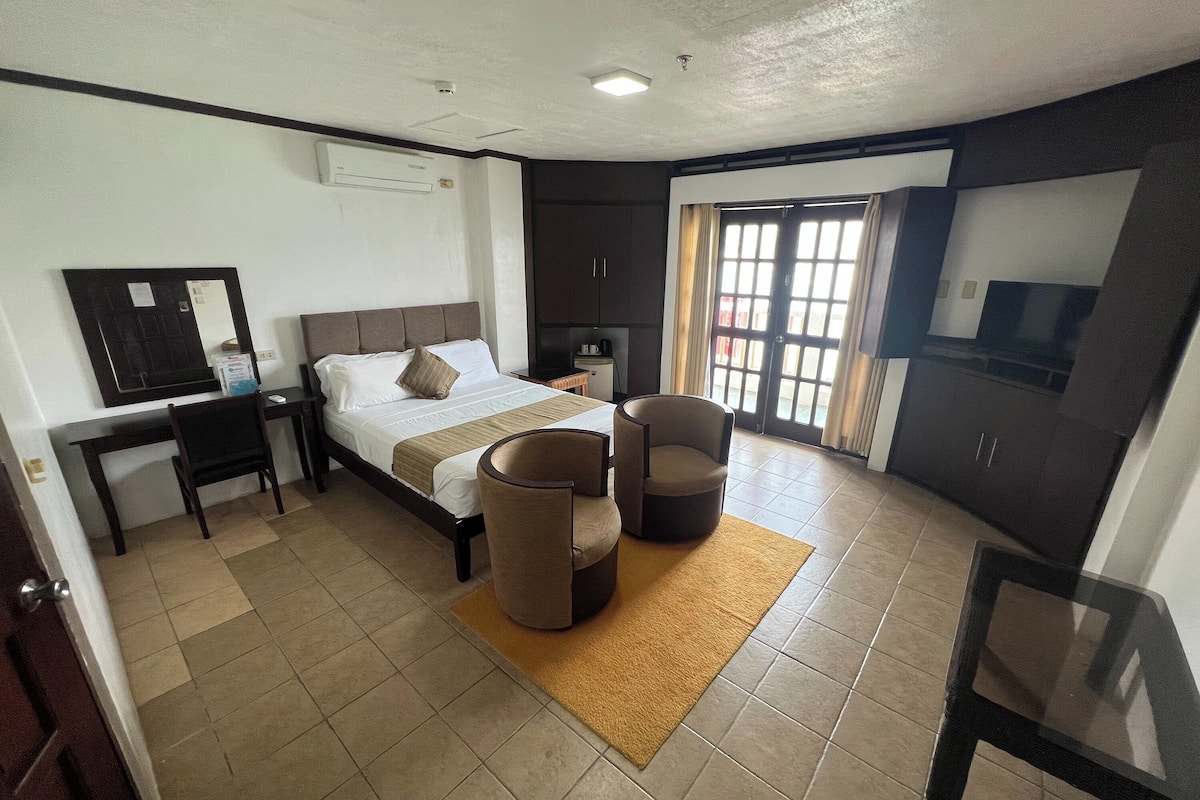 A hotel room with a bed, two armchairs, a desk, a TV, and a large window with curtains.