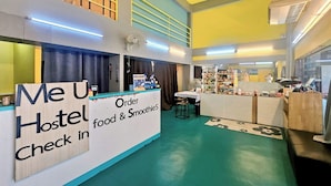 A hostel interior with a reception desk labeled "Me U Hostel Check in," a food and smoothies counter, and a seating area with a table and chairs. - Me U Hostel (Chiang Mai)