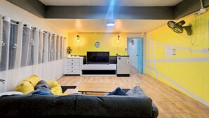 A living room with a sofa, a TV, and a small table. There is a clock on the wall and a fan on the wall. - Me U Hostel (Chiang Mai)