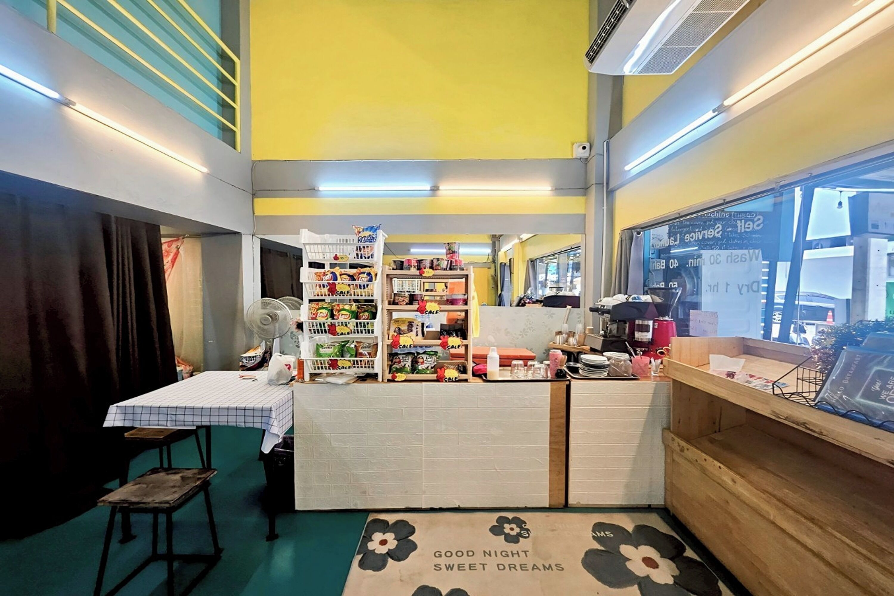A small, brightly lit room with a checkered table, a fan, and shelves stocked with snacks and drinks.