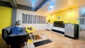 A modern living room with a sofa, coffee table, and a small kitchen area with a refrigerator and a TV. - Me U Hostel (Chiang Mai)