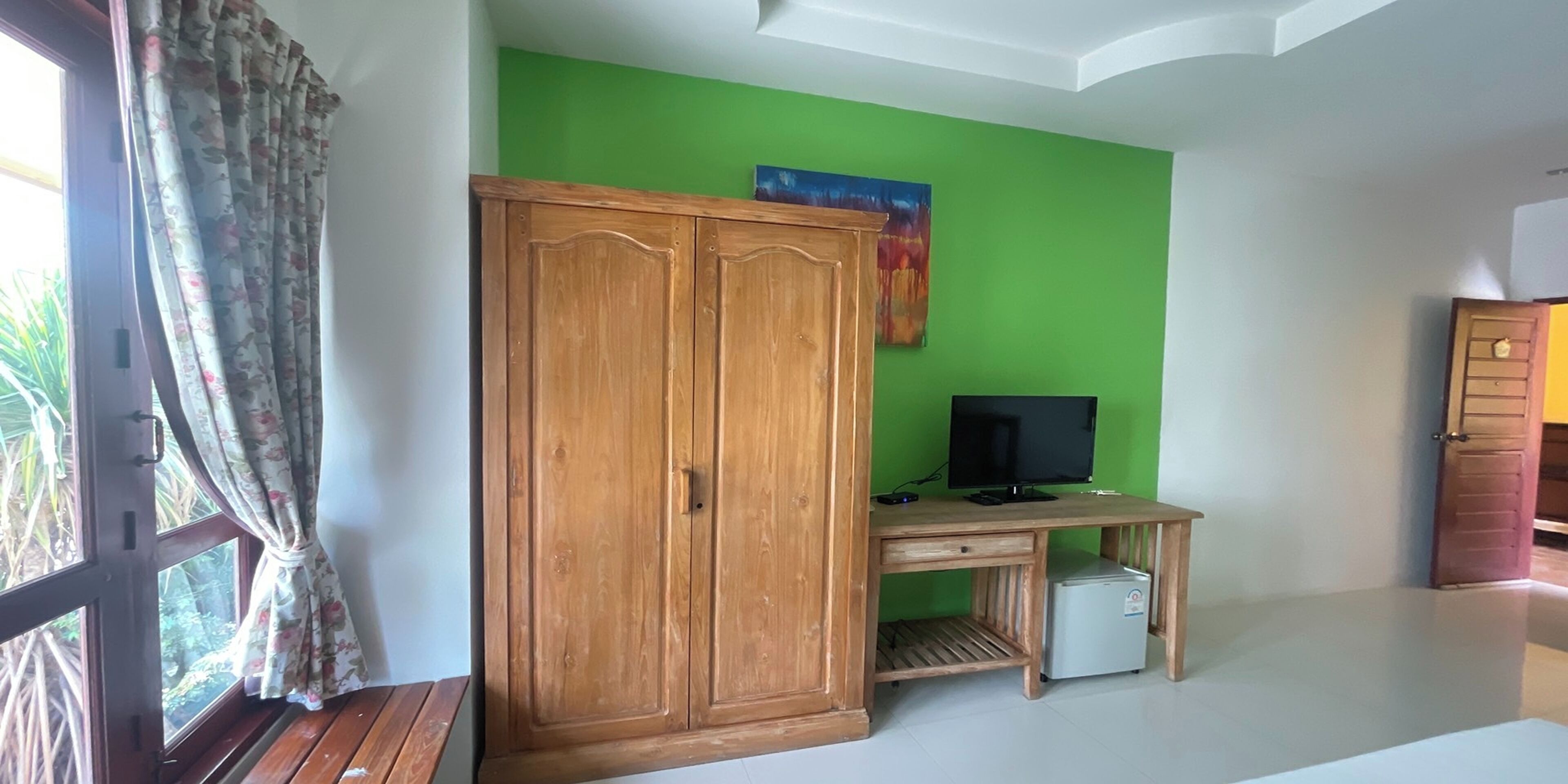 Una habitación con un armario de madera, un escritorio con un ordenador y un pequeño frigorífico, una pared verde y una ventana con cortinas.