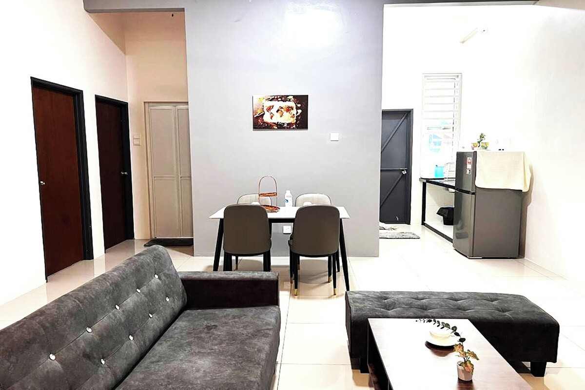 A modern living room with a grey sofa, a dining table with chairs, and a kitchen area with a refrigerator and a small table.