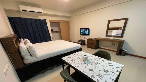 Ein kleiner, gut beleuchteter Raum mit einem Bett, einem Schreibtisch mit Computer, einem Fernseher und einem Spiegel.