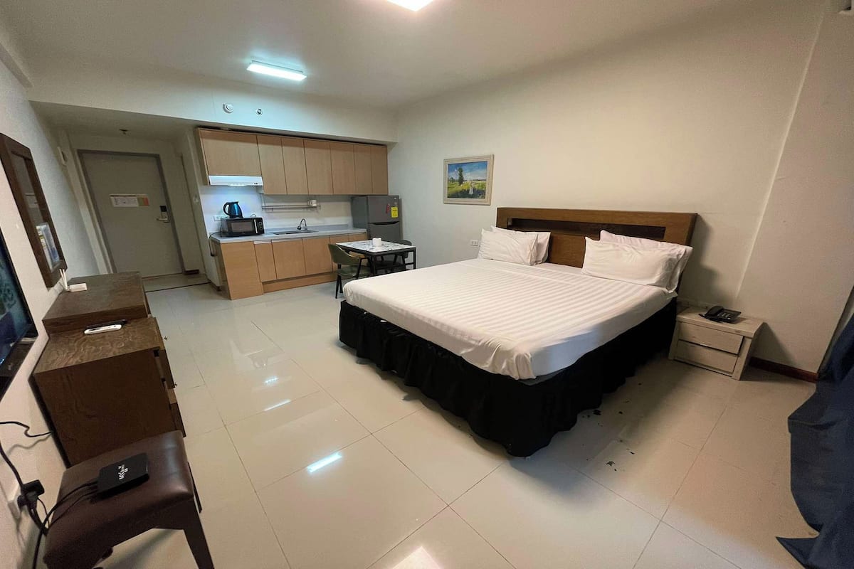 Una habitación de hotel con cama, escritorio, silla, mesita pequeña, mesita de noche, microondas, refrigerador y zona de cocina.