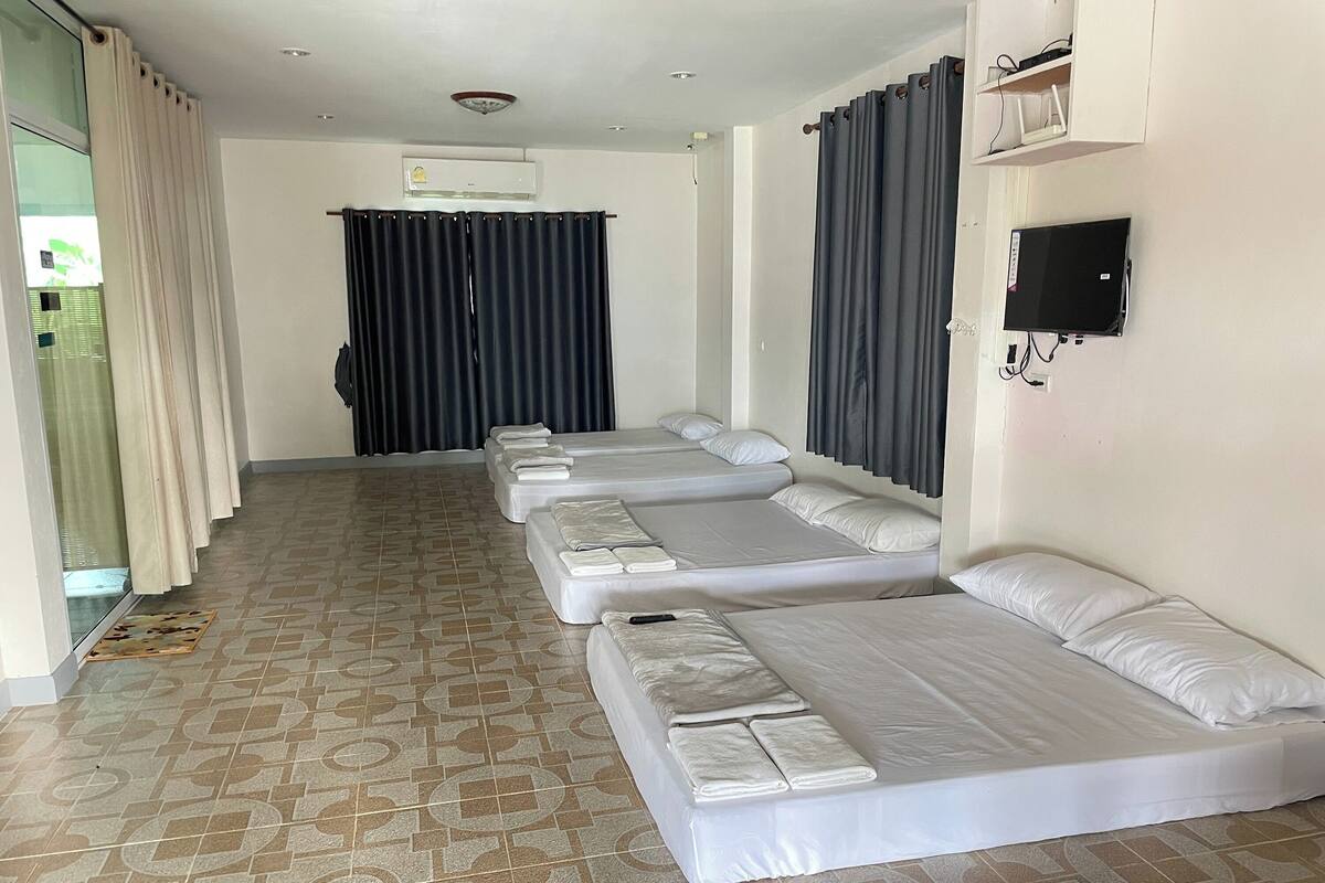 A hotel room with four single beds arranged in a row, a flat-screen TV mounted on the wall, and an air conditioning unit above the beds.