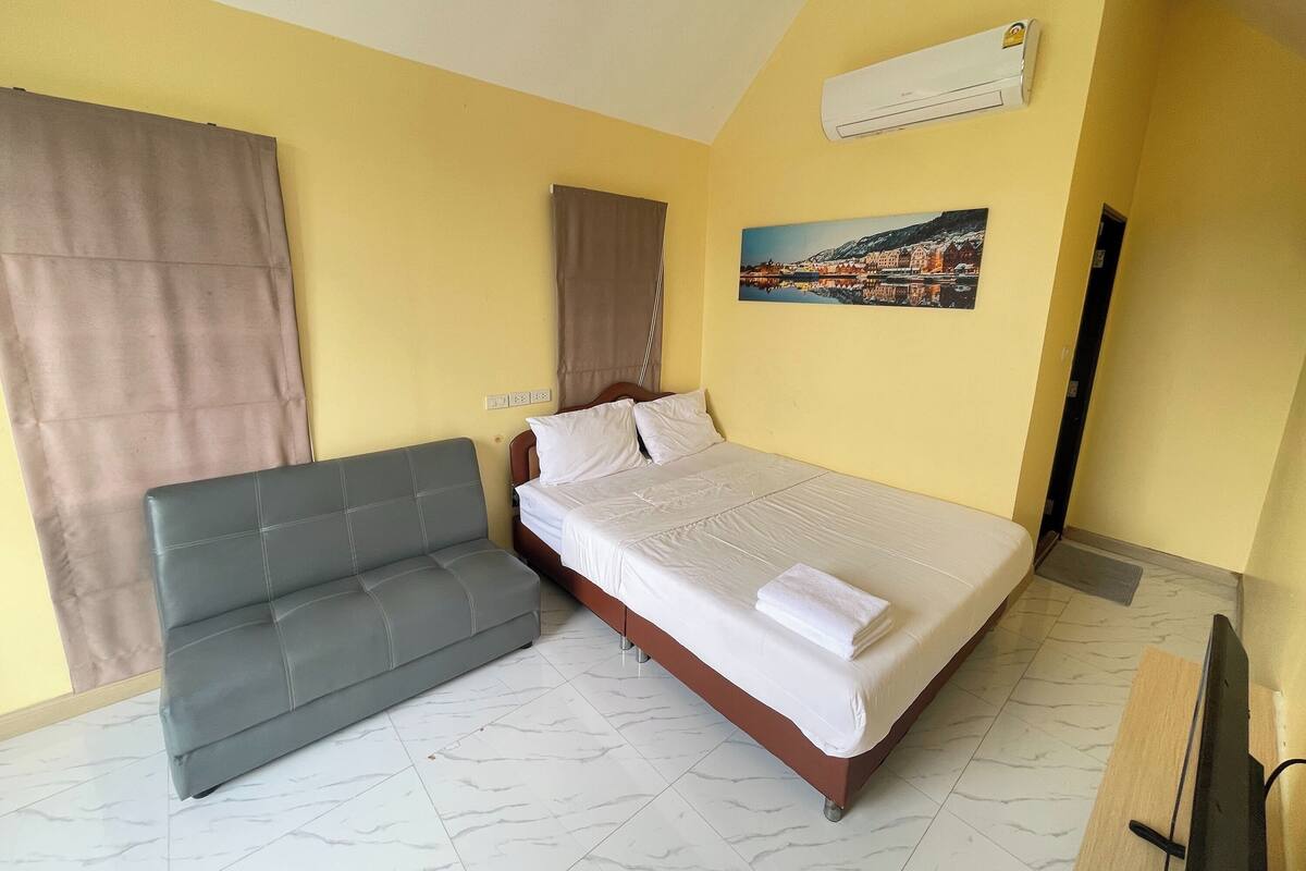 A hotel room with a bed, a grey sofa, a painting on the wall, and an air conditioning unit.