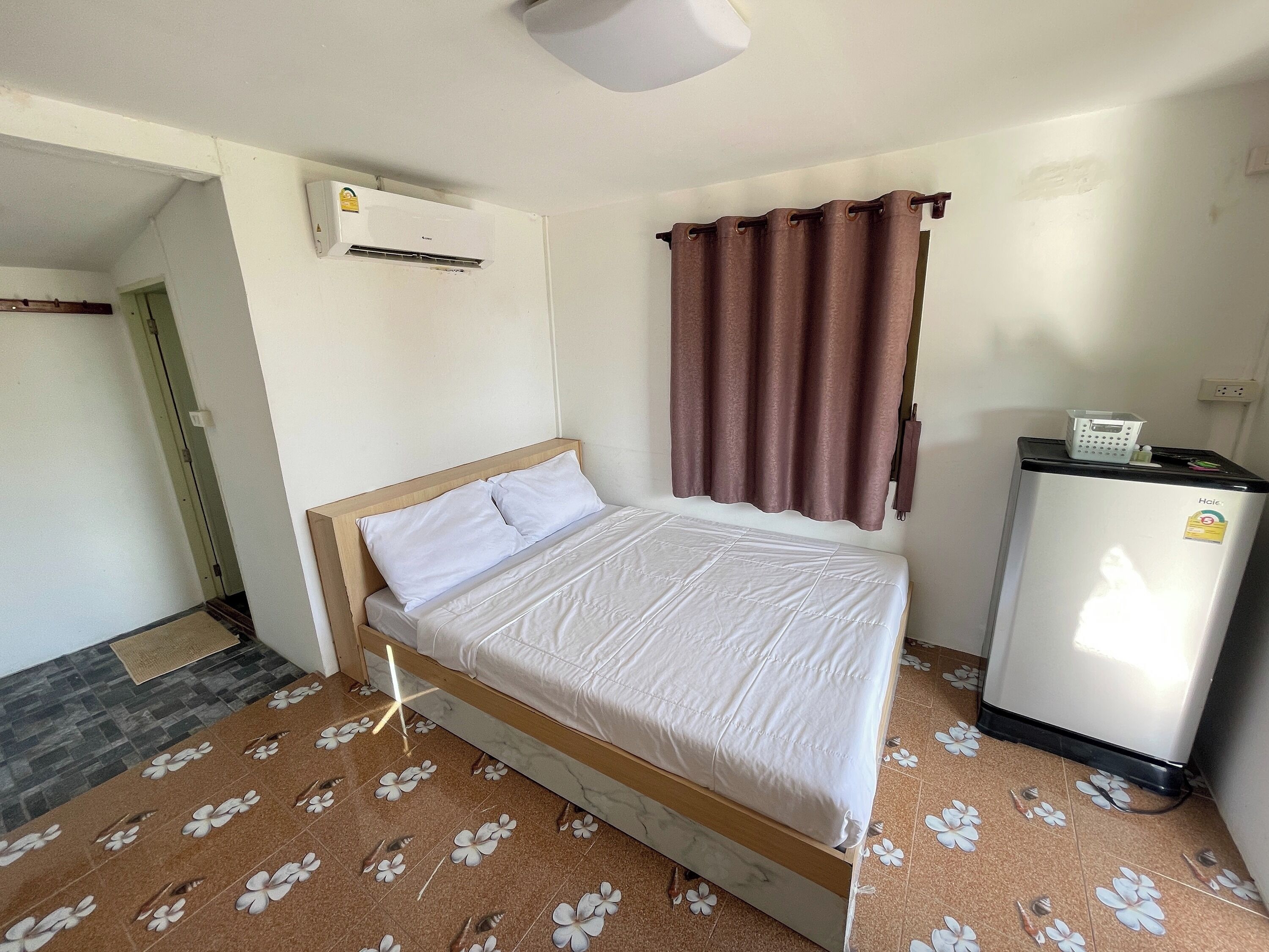 Una cama individual con estructura de madera y ropa de cama blanca, un refrigerador pequeño, un aire acondicionado instalado en la pared y una ventana con cortinas marrones.