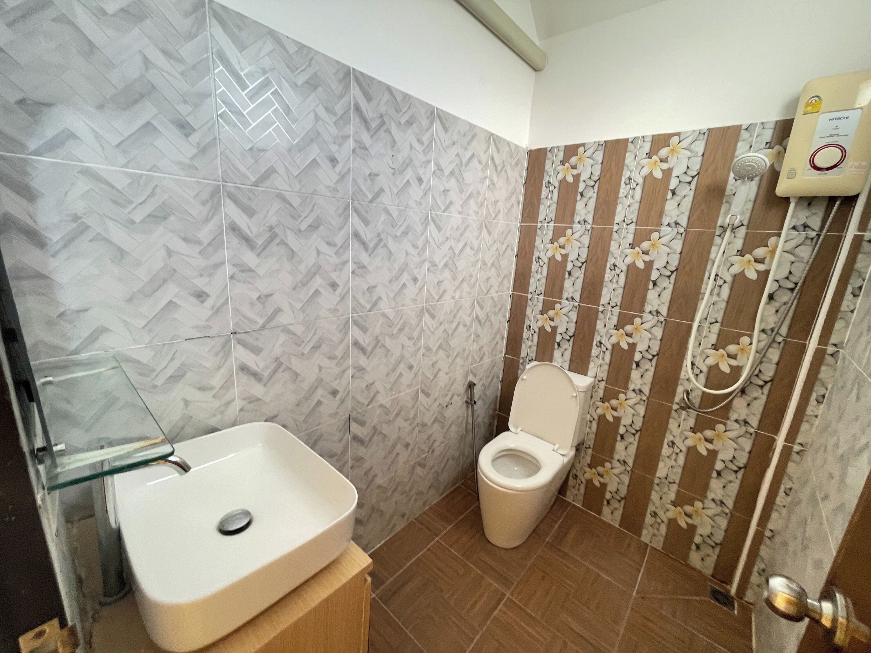 A bathroom with a herringbone tile pattern, a toilet, a shower with a handheld showerhead, and a glass-enclosed sink.