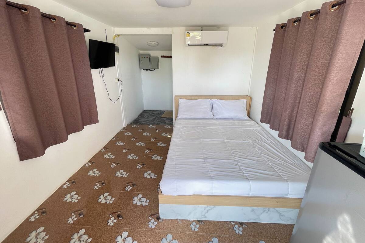 A small, single-room hotel room with a bed, a flat-screen TV mounted on the wall, an air conditioning unit, and a small kitchenette area.