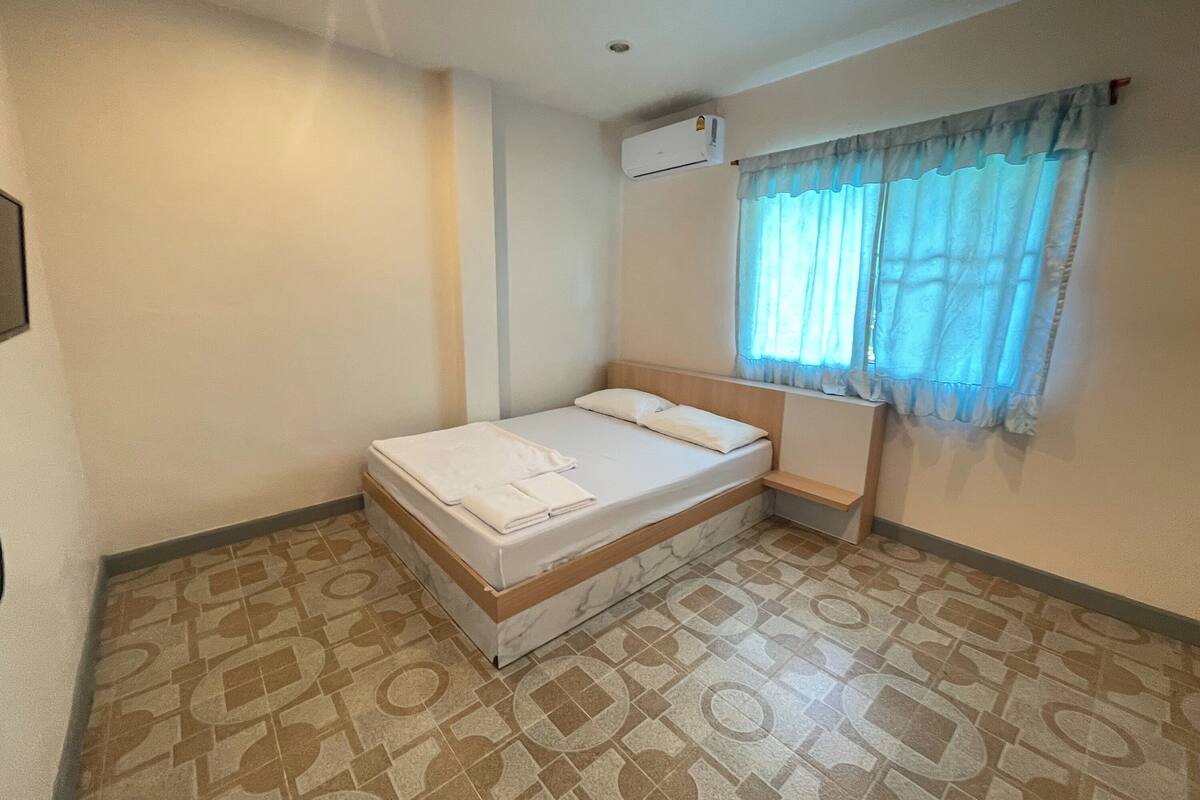 A small, neatly made bed with a wooden headboard and a white mattress, a window with light blue curtains, and an air conditioning unit on the wall.