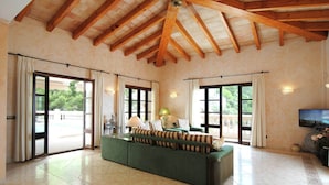 A spacious living room with a green sofa, wooden beams on the ceiling, large windows, and a television. - Villa in Canyamel with Bay Views and Pool (Canyamel)