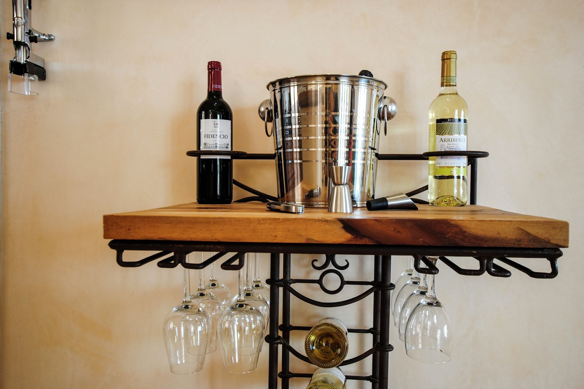 A wooden table with two wine bottles and a large metal container, possibly for ice, with several wine glasses hanging below.