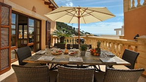 A dining table set with plates, glasses, and a large umbrella on a balcony overlooking a scenic view. - Villa in Canyamel with Bay Views and Pool (Canyamel)