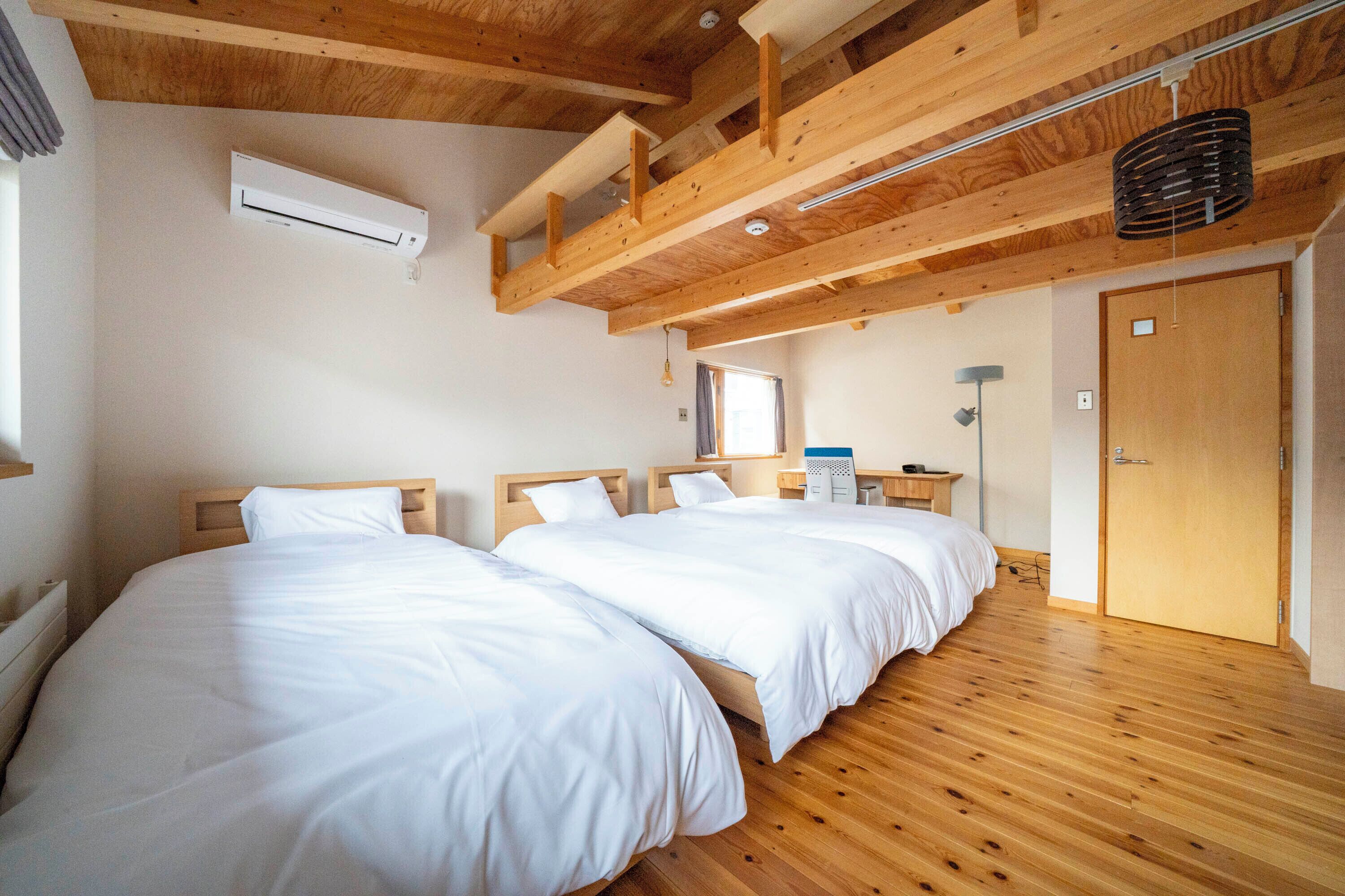 Quarto com duas camas, piso de madeira, ventoinha no teto e uma porta.
