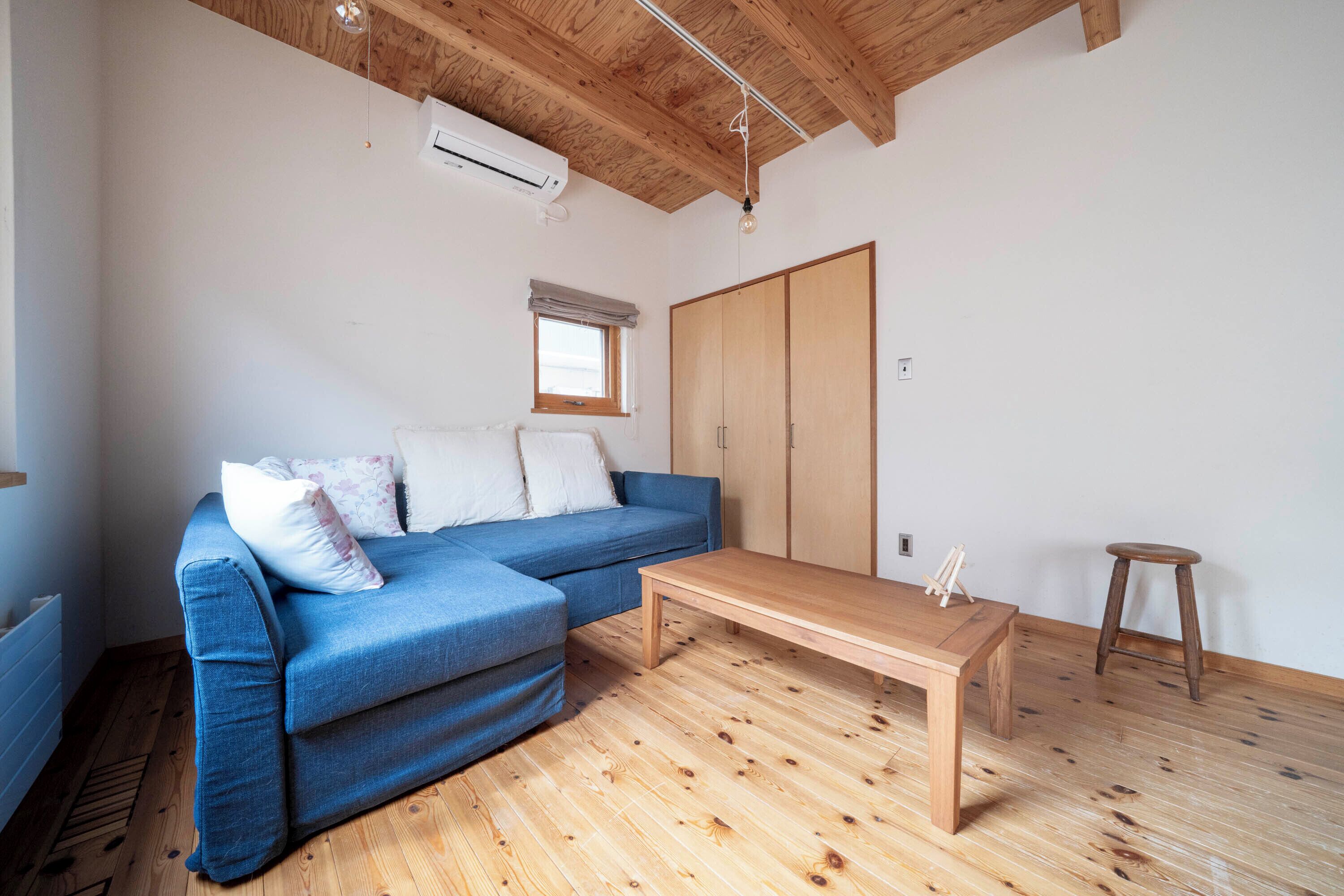A room with a blue sofa, wooden floor, a wooden table, a stool, a wardrobe, and a window with a curtain.