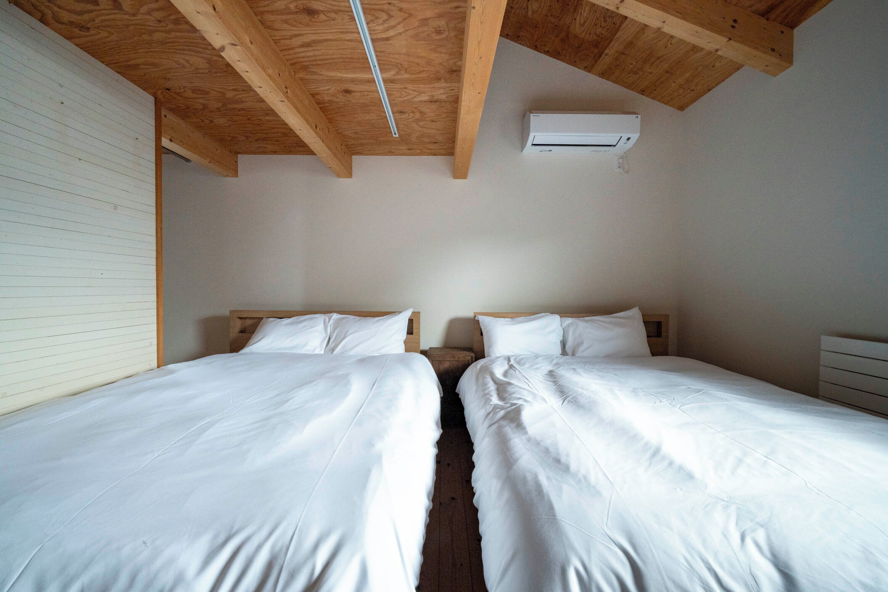 Quarto com duas camas, vigas de madeira no teto, um ar condicionado branco e uma parede branca.