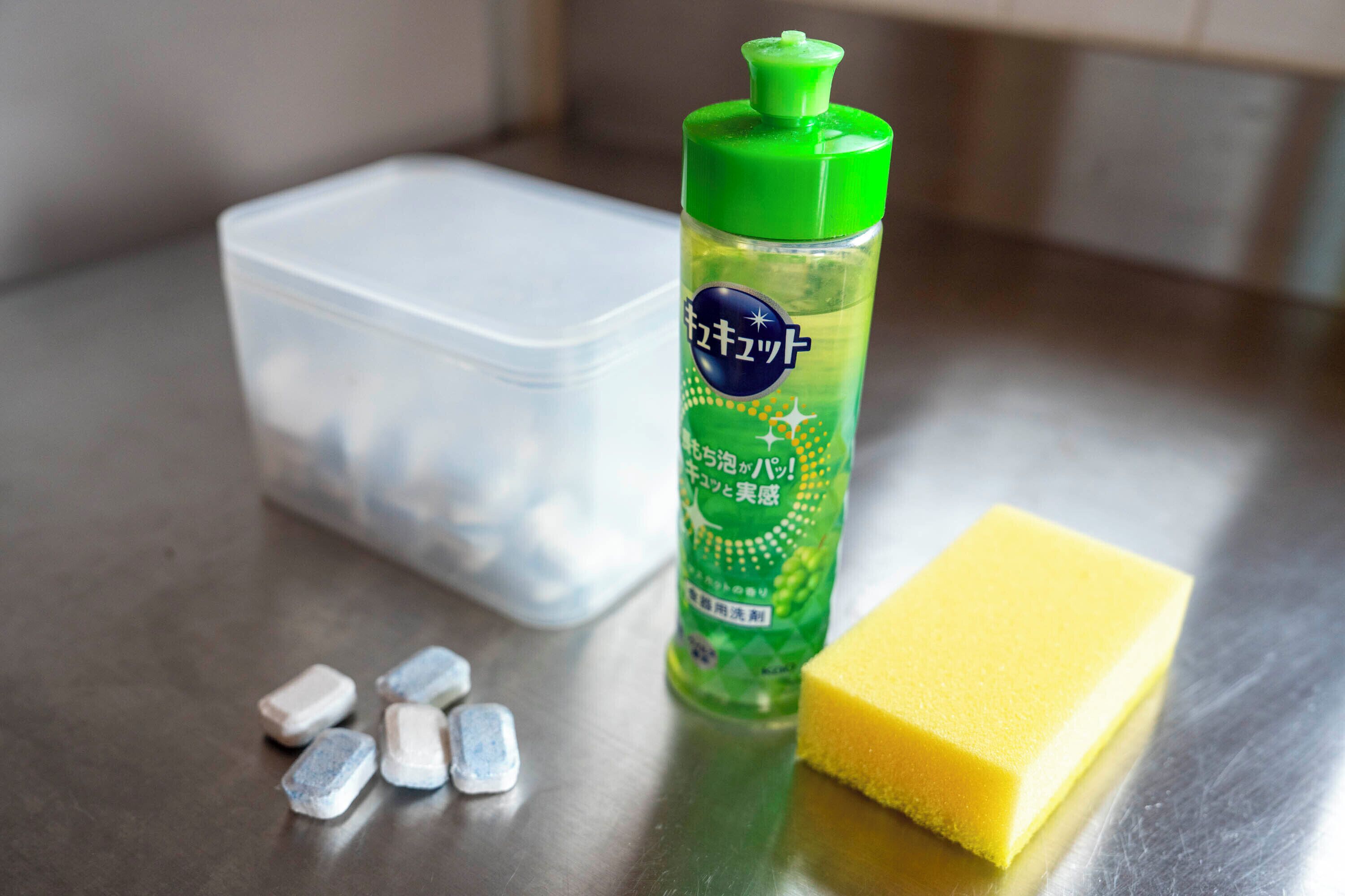 Un récipient en plastique, une bouteille verte avec une étiquette et une éponge jaune sur une surface métallique.
