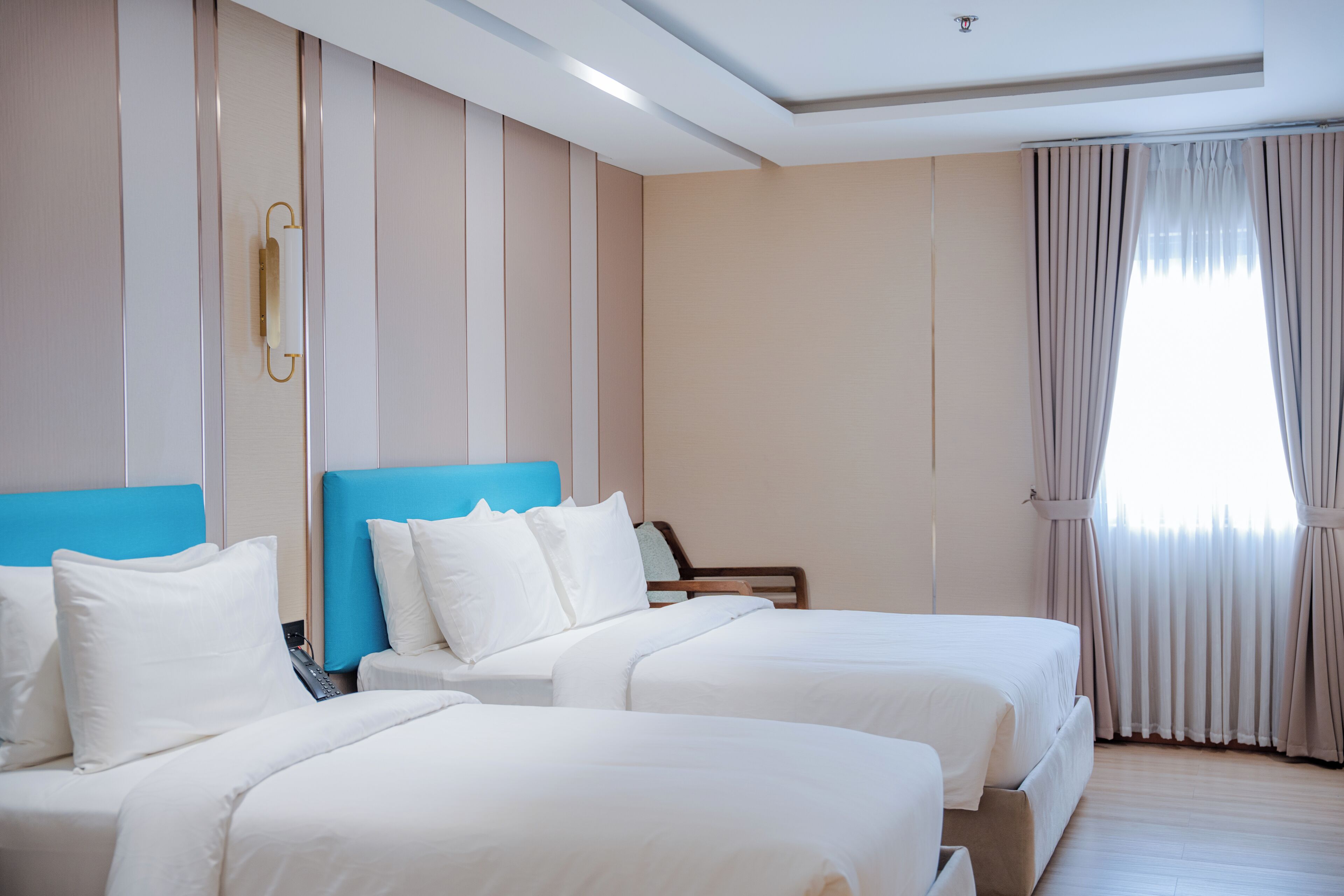 Ein Hotelzimmer mit zwei Betten, einem blauen Kopfteil und einem großen Fenster mit Vorhängen.