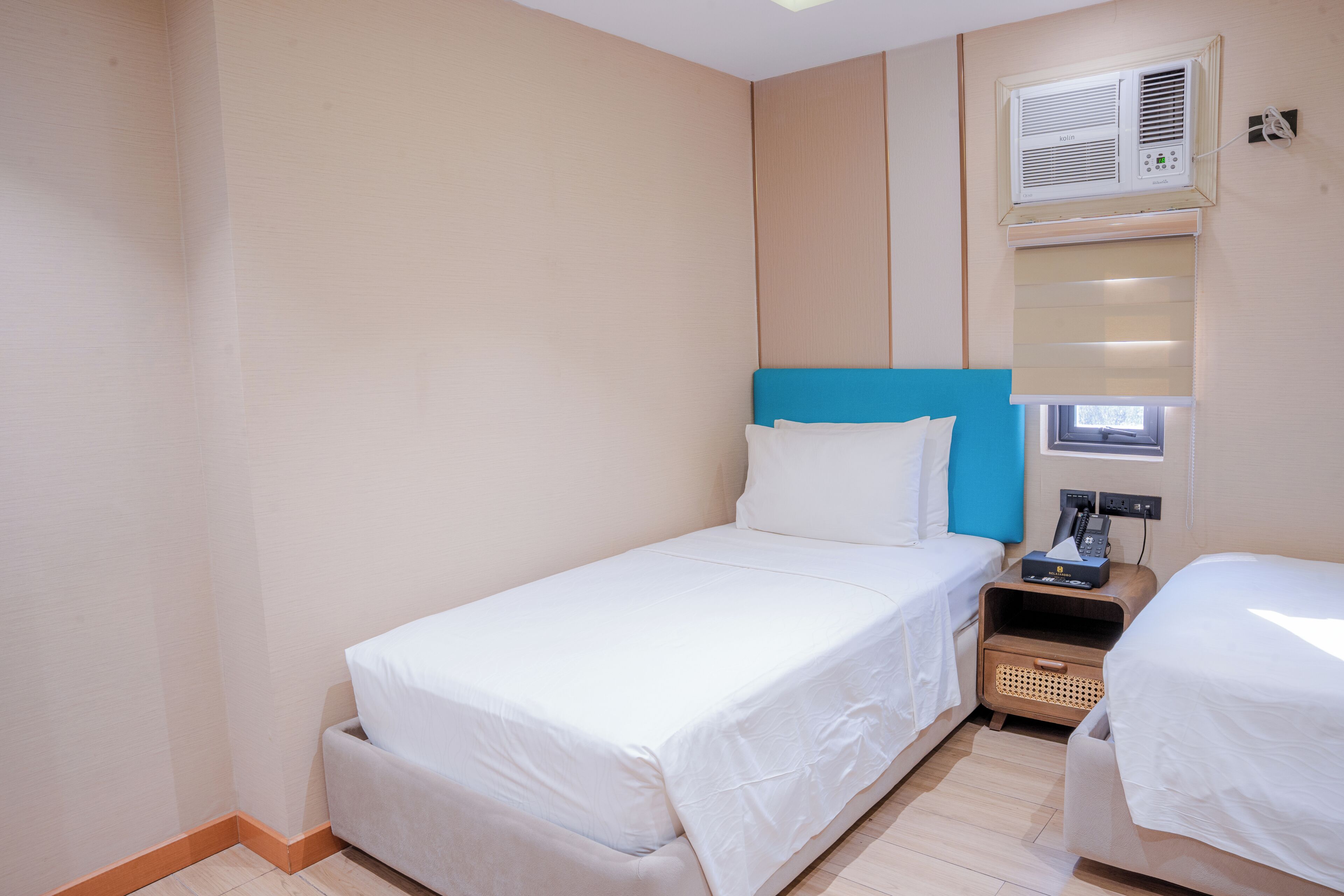 A hotel room with a single bed, a bedside table with a lamp, a small bedside table with a phone, and a wall-mounted air conditioner.