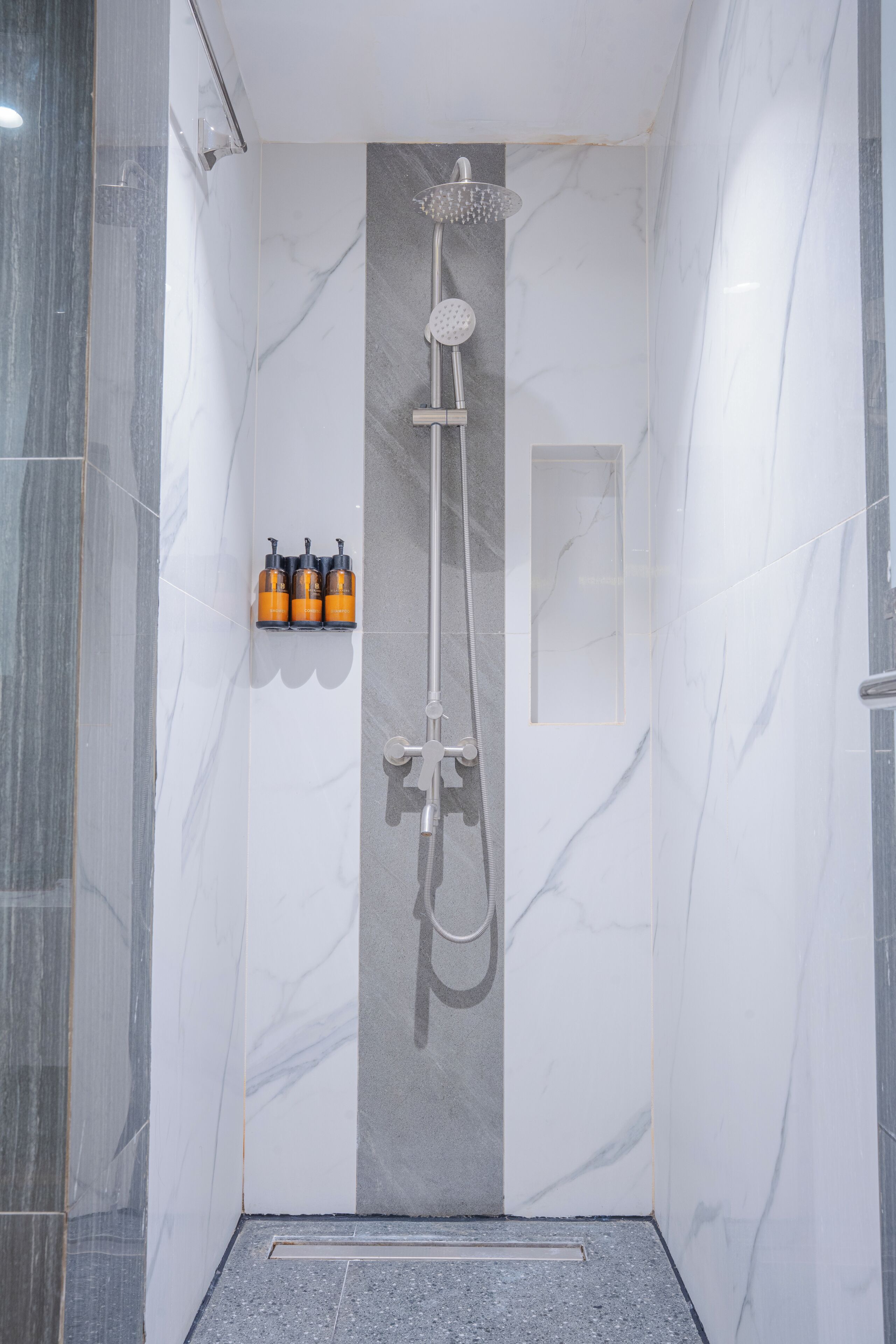 A modern shower with a handheld showerhead and a rain shower head, a small shelf with toiletries, and a marble wall with a vertical strip design.