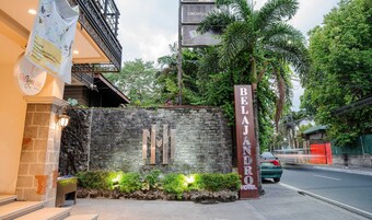 A hotel entrance with a stone wall, a sign for 'BELAJ ANDRO HOTEL,' and a street view with a car passing by.